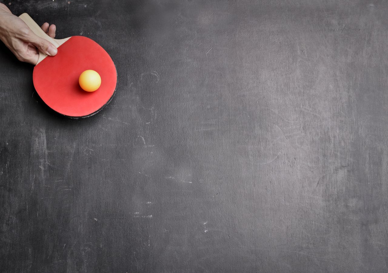 From above of crop male hand holding red ping pong racket and orange ball on gray tennis table